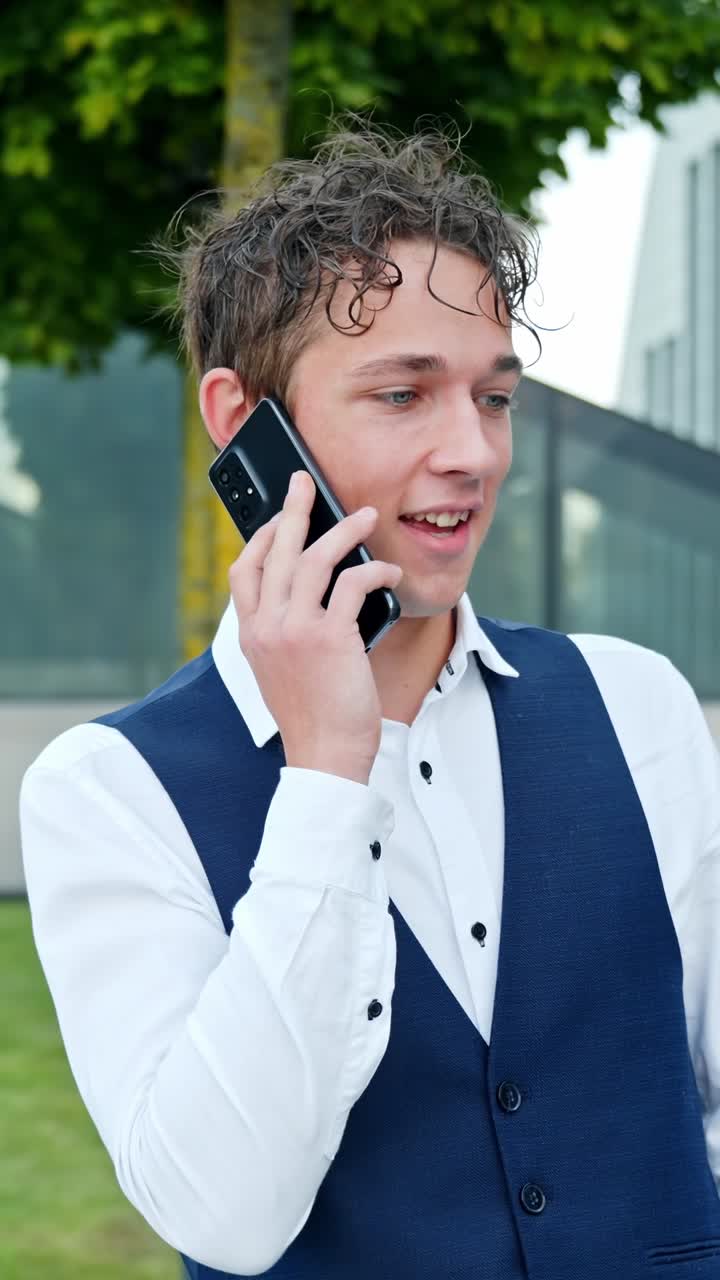 A vertical shot shows a young man in formal business attire speaking on his phone outdoors. He smiles, looks around, and appears happy during the animated conversation