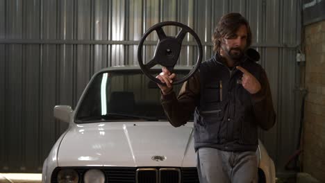 mechanic man holds up replacement steering wheel and points sitting on white car