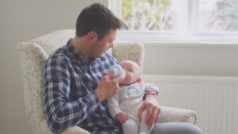 Father-feeding-baby-son-with-bottle-sitting-in-chair-at-home-together---shot-in-low-motion