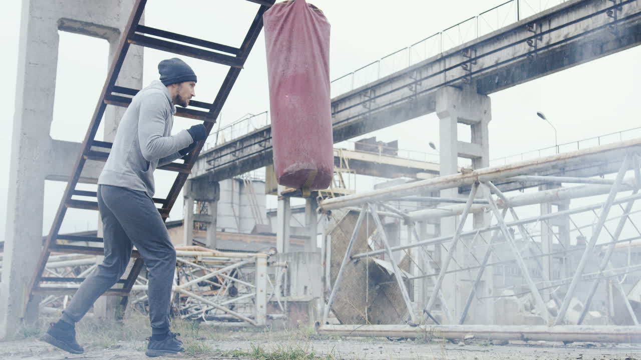hombre caucásico con gorro gris y ropa deportiva golpeando un saco de boxeo al aire libre en una fábrica abandonada en una mañana nublada