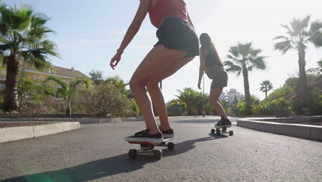 Zwei-Mädchen-Auf-Skateboards-In-Kurzen-Shorts-Fahren-In-Zeitlupe-Die-Straße-Entlang-Am-Strand-Und-An-Palmen-Entlang