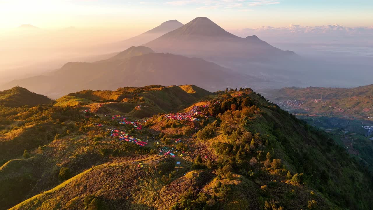 Vista aerea di tende colorate che punteggiano la cima erbosa della montagna con un'alba epica e imponenti montagne e dolci colline sullo sfondo. Paesaggio della cima del Monte Prau, Indonesia.