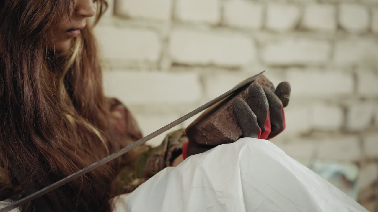 Primer plano de un niño marginado con pelo largo y guantes afilando una hoja sobre una piedra; el borde metálico raspa la superficie rugosa, expresión concentrada sobre una pared de ladrillo, ambiente áspero