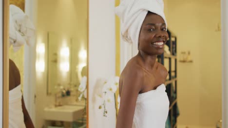 Portrait-of-smiling-african-american-woman-next-to-mirror-with-towel-in-bathroom