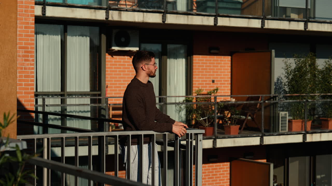 A young man steps onto a bright balcony, soaking up the sun and admiring the city view. Relaxed lifestyle moment in a modern apartment block on a beautiful sunny day