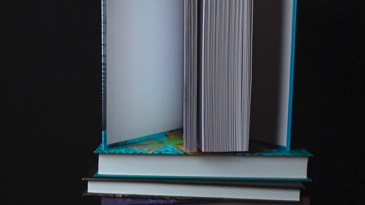 Close up or closeup footage that moves up slowly of hardcover colorful books in a pile horizontally while one on top is opened and is standing vertically on a dark black studio background. White pages
