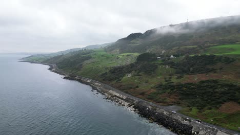 Toma-Panorámica-Aérea-Sobre-El-Hermoso-Paisaje-Rocoso-En-La-Costa-De-La-Carretera-Costera-De-Irlanda-Del-Norte-Cerca-De-La-Ciudad-De-Glenarm-Con-Vistas-A-La-Carretera-Rocosa,-El-Mar-Azul-Y-La-Montaña-En-Ascenso-Por-La-Mañana