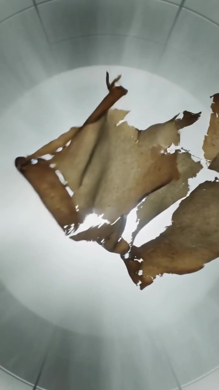 Vertikal video: Roterende brunt papir, der snor sig og mister fragmenter over skarpt lys i et kammer