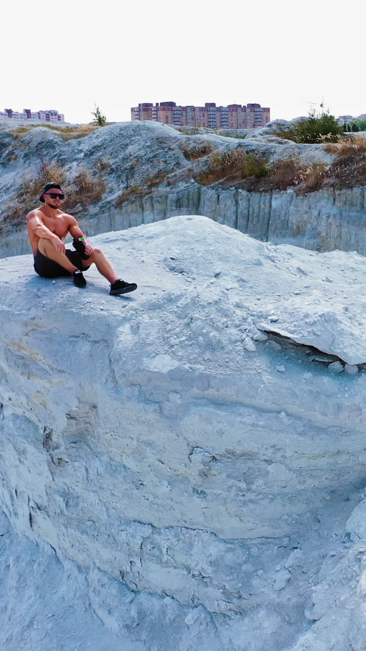 Brutal strong bodybuilder posing outdoor. Photoshoot in a quarry. Outdoor sports concept. Aerial view. Handsome strongman sits on a peak of a quarry. White landscape. Vertical video