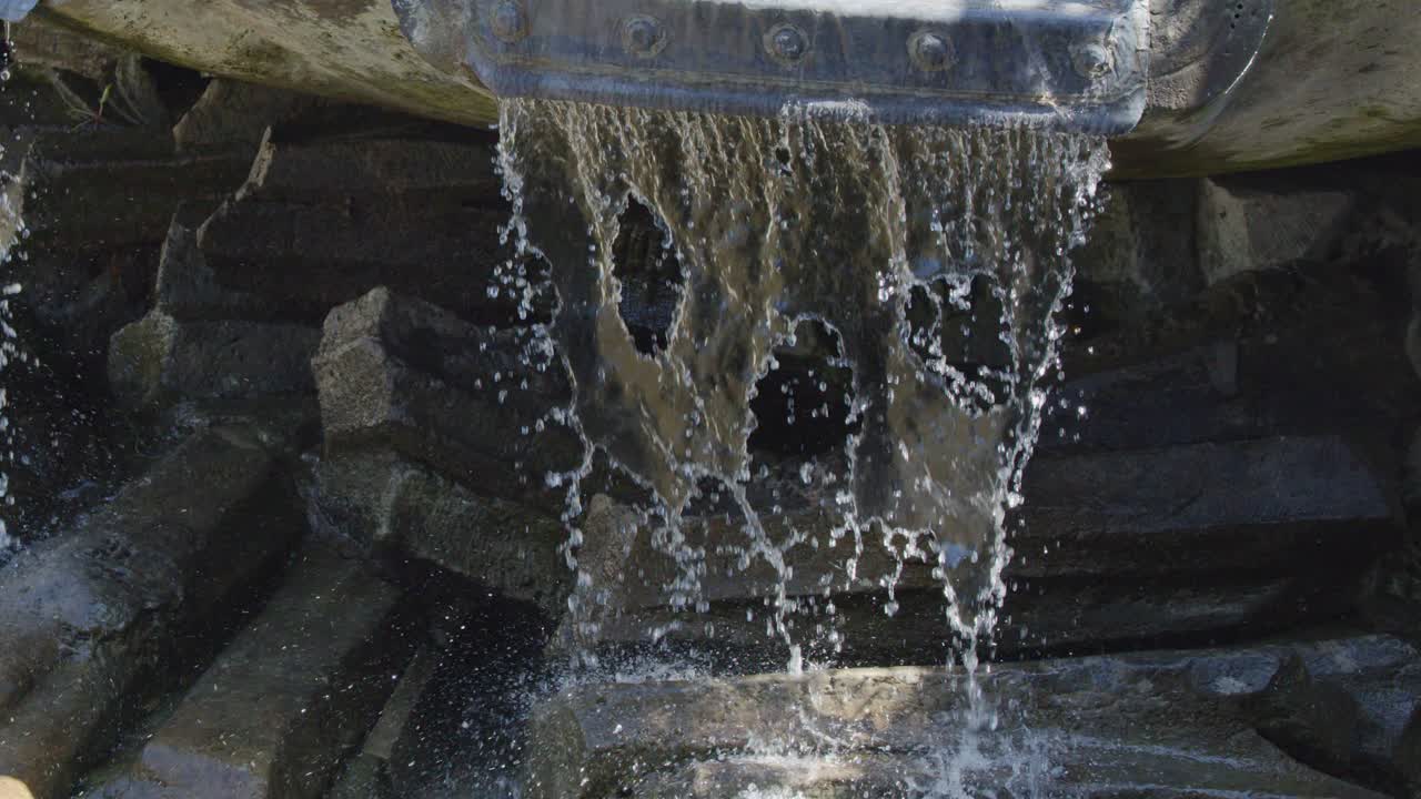 Water cascades from a sculpted stone fountain over dark rocks, sparkling in natural daylight