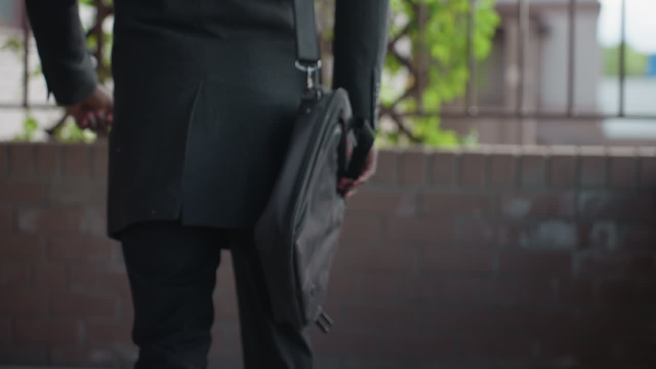 tight closeup shows strap clip of laptop bag swaying against dark coat, metal hook and ring bouncing during walk, minimal background softly blurred, focus on hardware movement and fabric texture