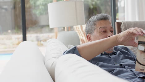 Feliz-Hombre-Birracial-Mayor-Relajándose-En-El-Sofá-Leyendo-Un-Libro-En-La-Sala-De-Estar,-Copiando-Espacio,-Cámara-Lenta