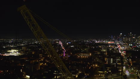 Toma-Aérea-De-Seguimiento-De-Una-Grúa-De-Construcción-Con-Fondo-De-Paisaje-Urbano-Iluminado-Por-La-Noche.