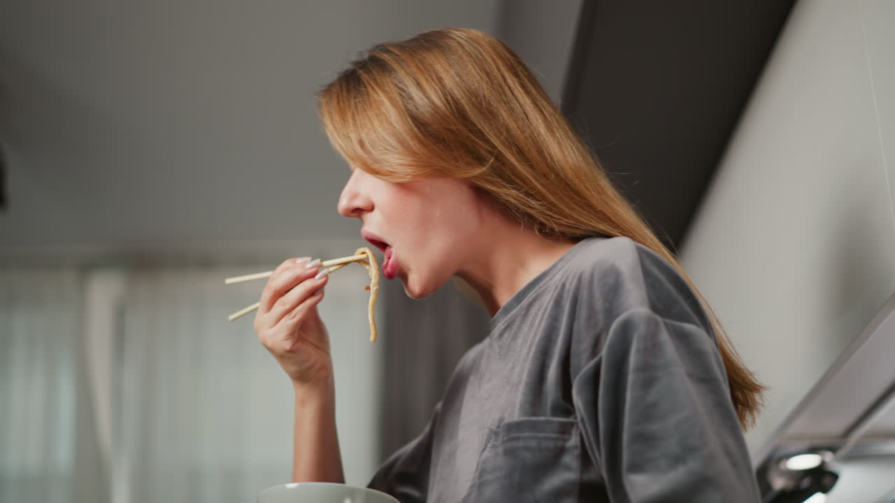 Side view of girl enjoying pasta while holding it with chopsticks, standing against soft blur background featuring sheer curtain and bright kitchen cabinet light