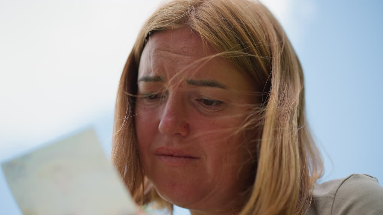 Close up of woman gazing sadly at photo in her hand under soft daylight sky, wind blowing strands of hair across face, emotion of grief, longing, and reflection visible through expressive eyes