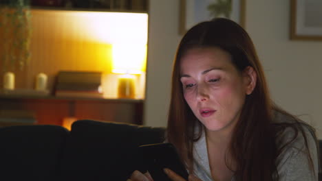 woman sitting on sofa at home at night streaming or looking at online content on mobile phone 1