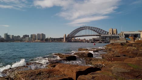 Calm-and-stunning-video-footage-of-Sydney-Harbour-Bridge-featuring-city-office-builidings,-ocean-water,-rocks,-heritage-buildings,-and-boat-passing-through-on-a-clear-sky-sunny-day