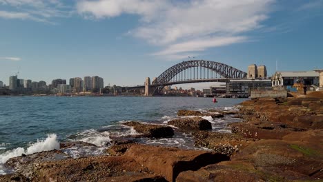 calm and stunning video footage of sydney harbour bridge featuring city office builidings, ocean water, rocks, heritage buildings, and boat passing through on a clear sky sunny day