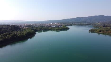 Panoramic-view-over-a-beautiful-landscape-with-a-natural-lake-surrounded-by-trees,-and-a-town-nearby-the-mountains,-Spain