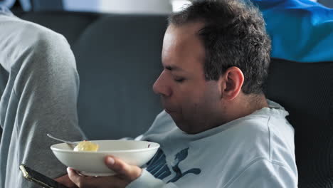 fotografía cinematográfica en primer plano de un hombre blanco de cabello oscuro sentado en un sofá negro comiendo mientras revisa su teléfono con las piernas cruzadas en su apartamento durante un día