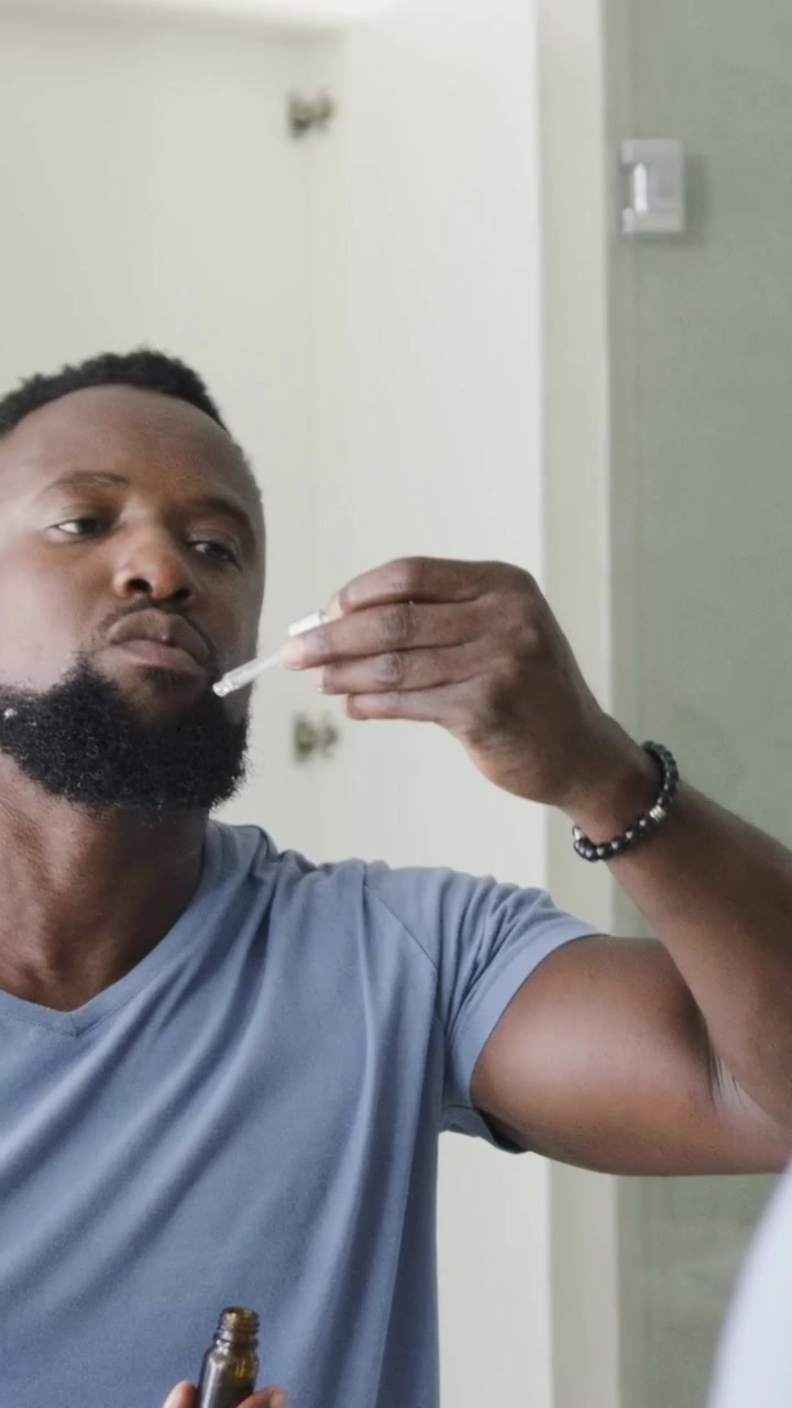 Vertical video: Touching face man in blue tee tilting and grooming beard with amber dropper at sink