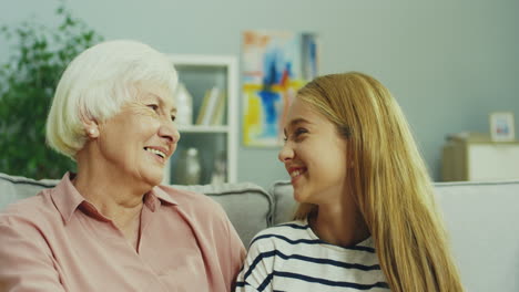 Portrait-Of-The-Teen-Pretty-Girl-And-Her-Senior-Grandmother-Looking-At-Each-Other,-Then-Turning-To-The-Camera-And-Smiling-On-The-Sofa-At-Home