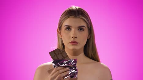 medium static shot of young attractive woman while she enjoys a bite of the vegan chocolate bar and looks with a satisfied saturated look to the camera in front of turquoise background in slow motion