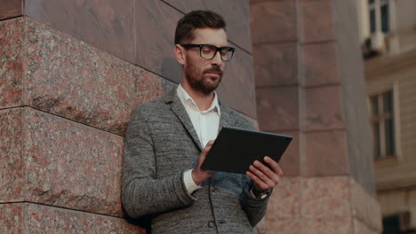 stylish businessman standing outdoor and leaning against the wall while tapping and scrolling on the tablet device