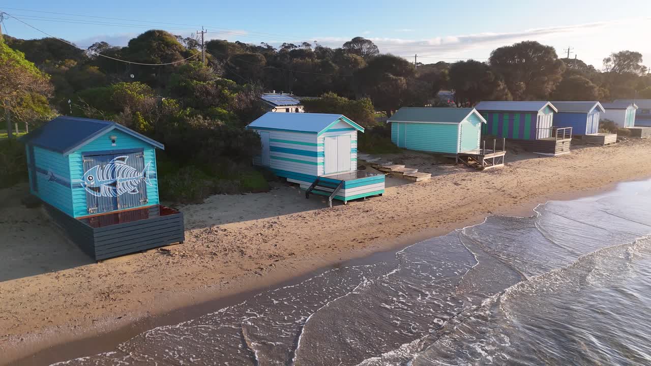 Drone glides over vibrant bathing boxes on sandy beach, gentle waves, soft morning sunlight