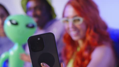 fotografía de estudio de un grupo de amigos de la generación z sentados en el sofá posando para una selfie con un alienígena de juguete en el teléfono móvil 5