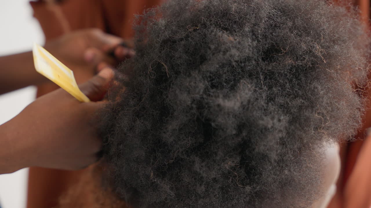 Mãos de cima modelando um cabelo afro preto com pente garfo e dedos, criando elevação e volume enquanto suaviza as bordas, luz de estúdio realça textura e cachos, interação amigável e cuidadosa, brincalhona, mas atenta