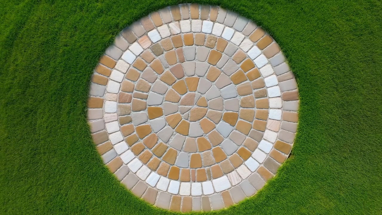 Orthogonal top-down view of full circular stone mosaic flush with trimmed lawn, showing concentric rings of irregular rounded rectangular and trapezoidal tiles in warm beige, tan, cream, and gray
