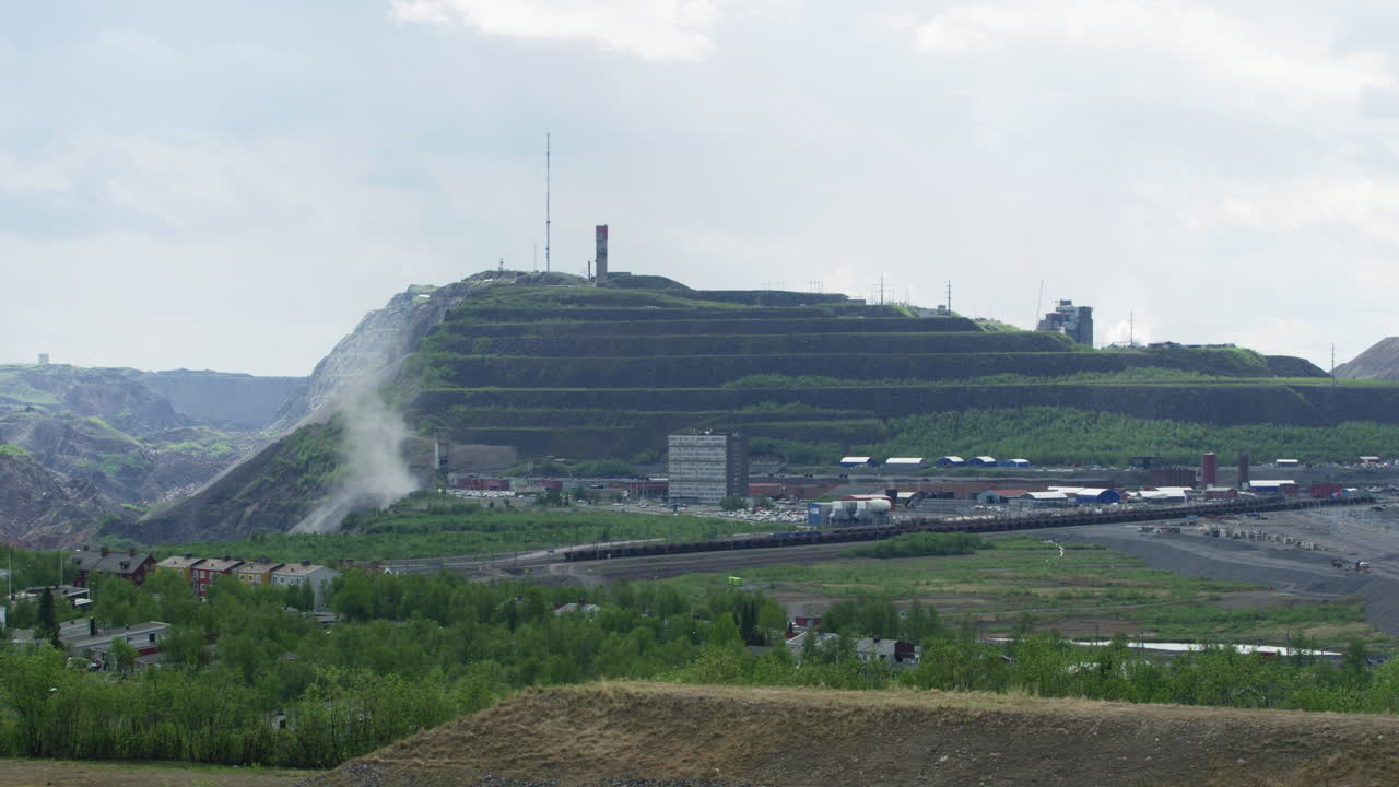 Panning across Kiruna town to the Kiruna iron ore mine run by Luossavaara-Kiirunavaara AB LKAB in Kiruna, Norrbotten, Lapland, Sweden. Card on road. Dust rising from the rocky talus slope