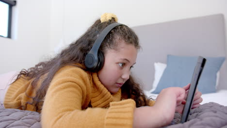 Happy-biracial-girl-lying-on-bed,-using-tablet-and-headphones-in-sunny-bedroom