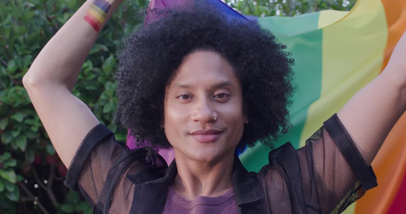 Uomo afroamericano che solleva una bandiera arcobaleno del Pride con le braccia davanti a della vegetazione, esprimendo orgoglio.