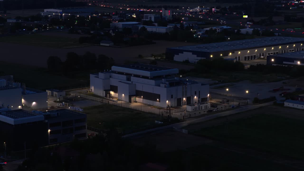 Aerial night view of a modern data center in Warsaw, Poland, surrounded by logistics hubs and industrial warehouses. Illuminated secure facility for servers, cloud storage, and digital networks