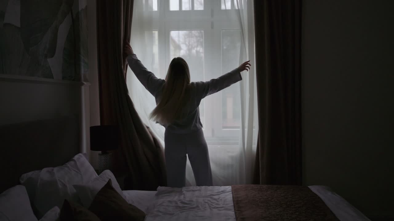 Una giovane donna si trova in una camera da letto e apre le tende per far entrare la soffusa luce del mattino