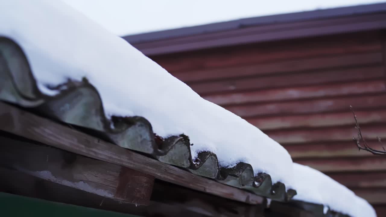 Snow piled on a corrugated metal eave beside a rustic wooden building, winter home exterior with frost and frozen runoff, rural preparedness and seasonal upkeep concept