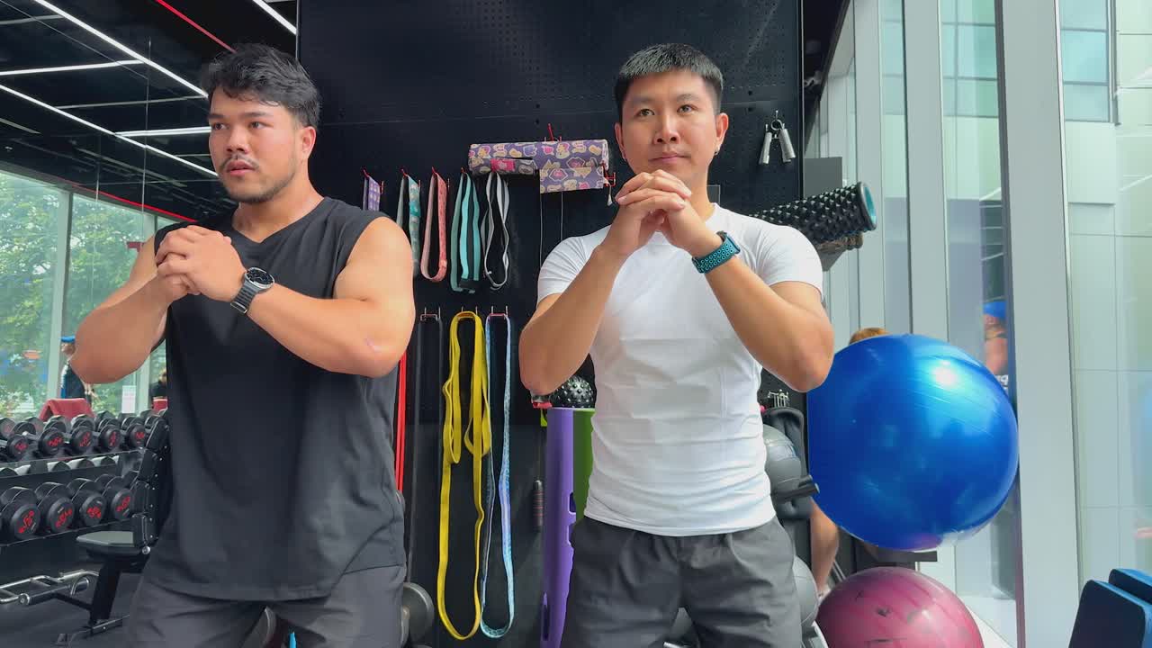 Two athletic men execute synchronized bodyweight squats in a brightly lit fitness center, surrounded by gym equipment and large windows overlooking greenery