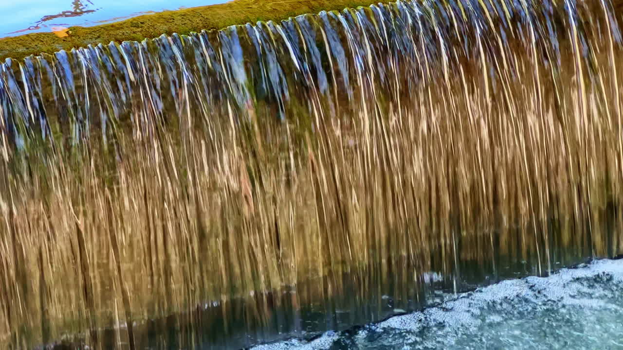Wasser fällt kaskadenartig einen Steinvorsprung hinab in ein klares Becken, das von üppigem Grün umgeben ist, im warmen Sonnenlicht eines friedlichen Nachmittags.