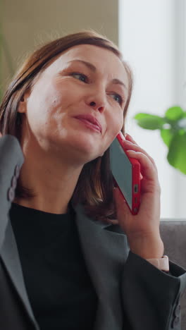 Smiling businesswoman in formal suit talking on smartphone while sitting comfortably on sofa in bright office lobby filled with green indoor plants and natural light coming through large window