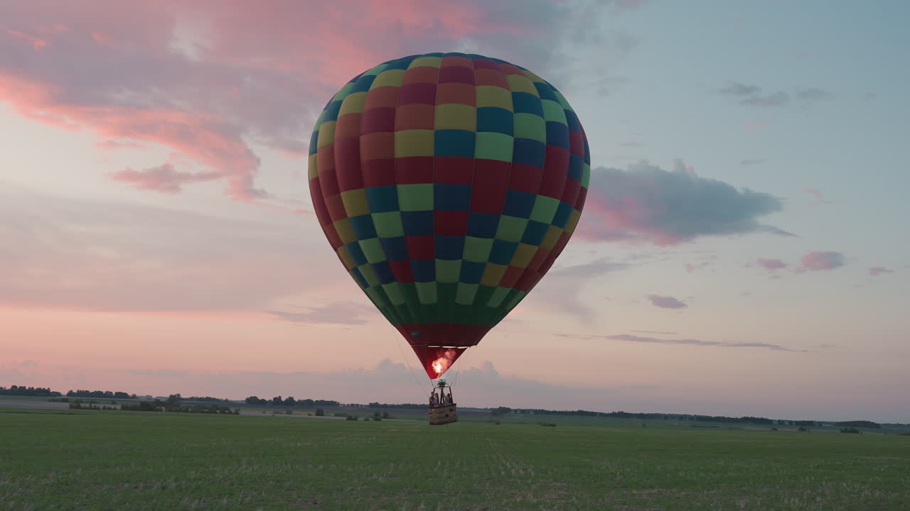 passengers in hot air balloon drifting low over green farmland at sunset burner flame flickering against pastel sky as distant village and tree line stretch across horizon