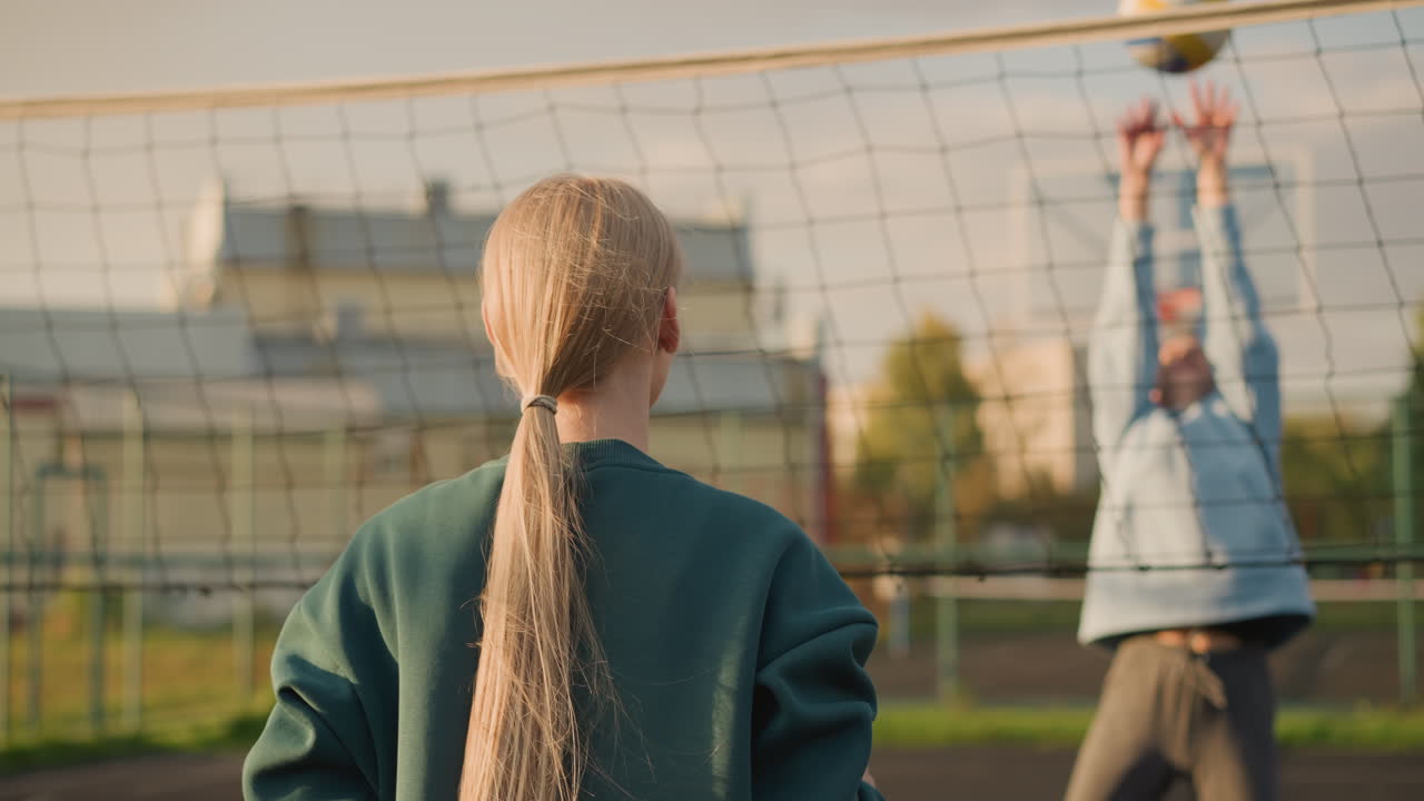 초록색 후디를 입은 여성이 야외에서 배구 경기를 하고 있으며, 배경에는 건물의 명확한 시야가 있으며, 배구 활동은 네트와 주변 개방된 공간으로 촬영되었습니다.