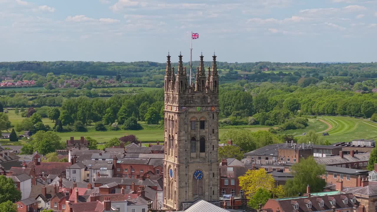 Umkreisende Aufnahme des Glockenturms der St. Mary’s Church in Warwick mit wehender Englandflagge an der Spitze, die die umliegende historische Stadt- und Dorflandschaft offenbart.