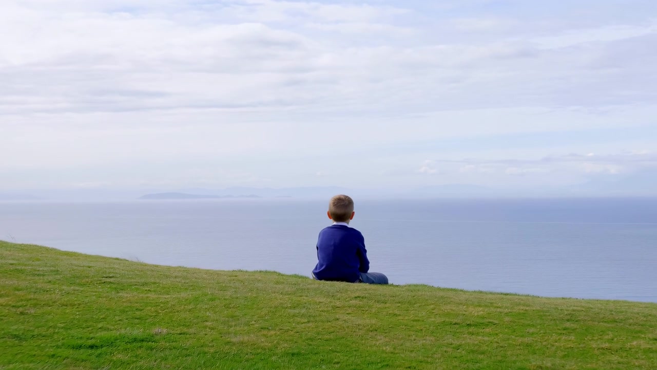 A lone person sits on a green grassy slope at center-right of the frame, facing away toward a pale, cloudy sky and a calm ocean horizon. Soft diffuse daylight