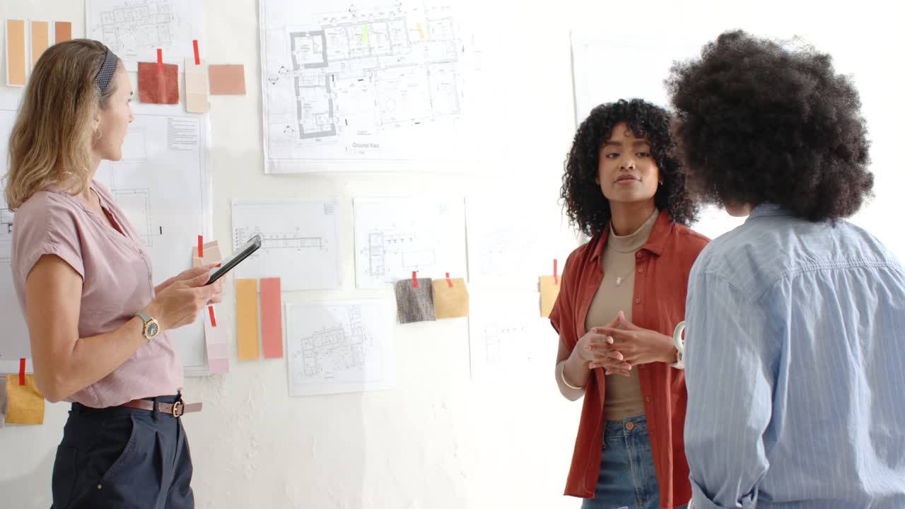 Diverse female team prompted by tablet, presenting, aligning on pinned plans to decide in studio