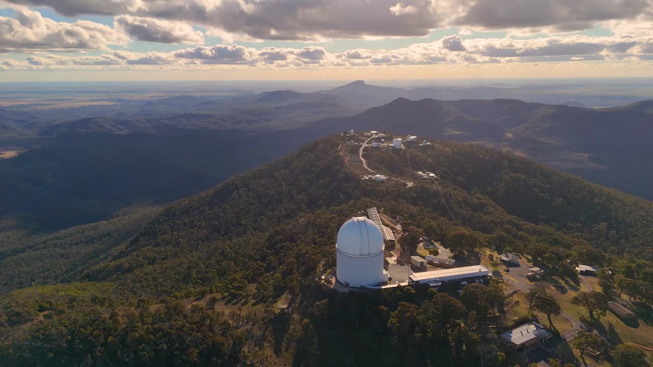 Sweeping drone footage reveals a large astronomical observatory atop a forested mountain ridge at sunset, surrounded by rugged terrain and dramatic skies
