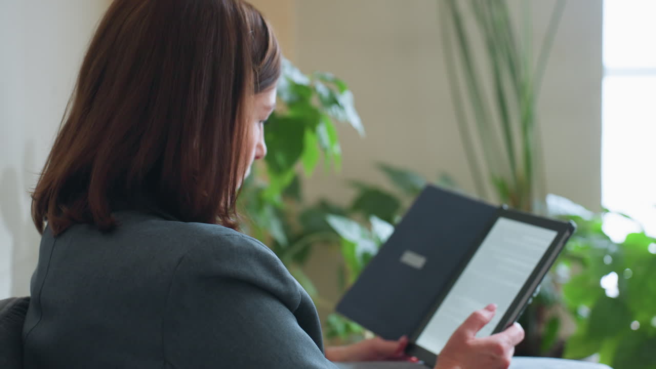Mature woman sitting indoors holding digital e-reader with blank screen while relaxing near window with natural light and surrounded by green houseplants in calm quiet home environment