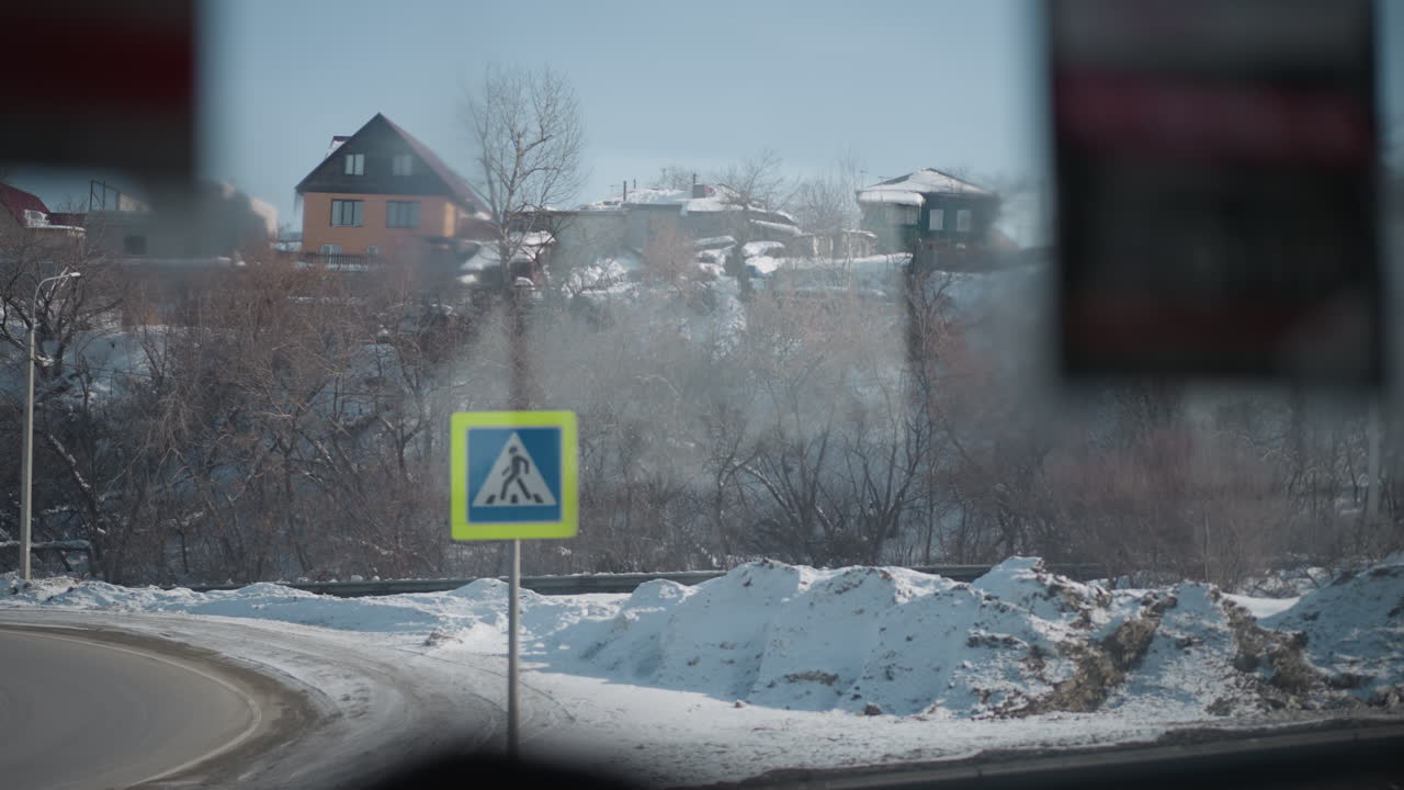 Window view from moving bus across winter city shows snowy hillside with houses, leafless trees, distant traffic light and sign posts, barrier along road, calm daylight and slow motion