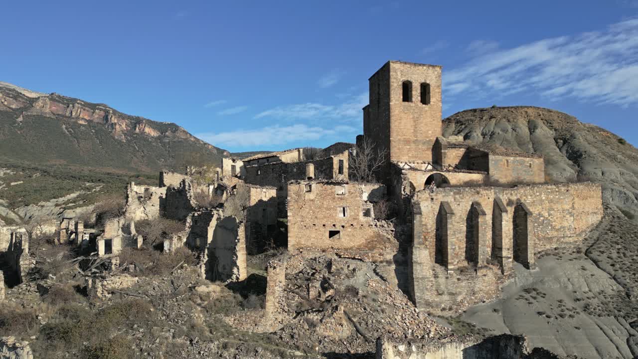 Vista aerea delle rovine medievali di Esco, una città fantasma abbandonata su un fianco di una collina in Aragona, Spagna, con la sua antica torre campanaria e case in pietra fatiscenti sotto un cielo azzurro e limpido